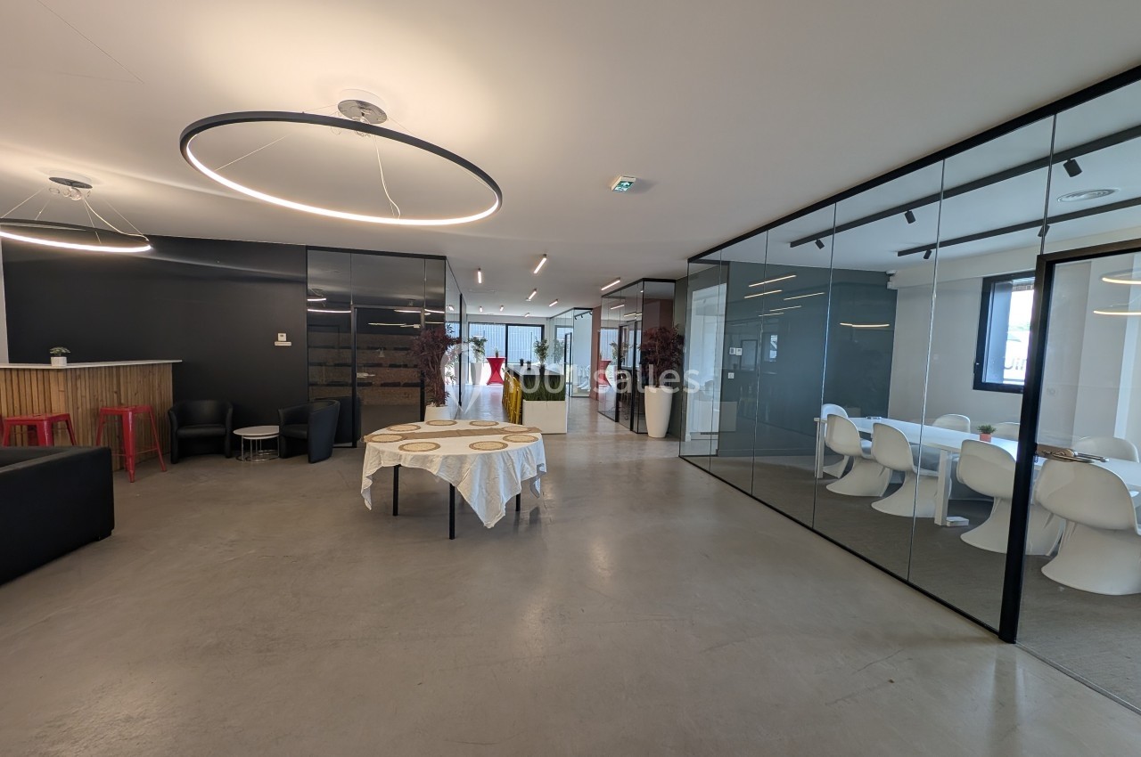 Espace intérieur moderne avec table ronde au centre, coin salon à gauche et salle de réunion vitrée à droite.