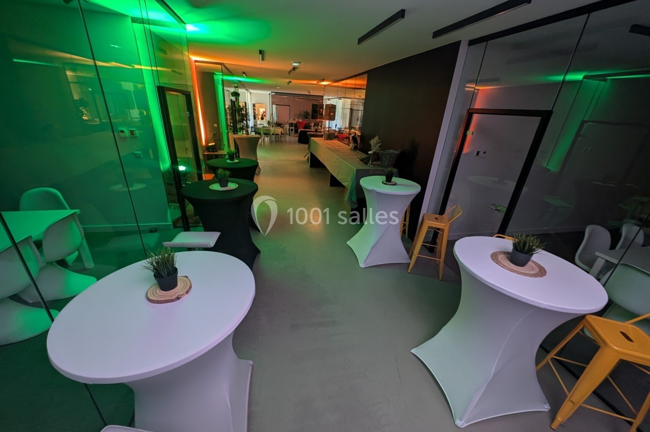 Salle moderne avec tables hautes blanches, éclairage coloré et murs vitrés, prête pour un événement.