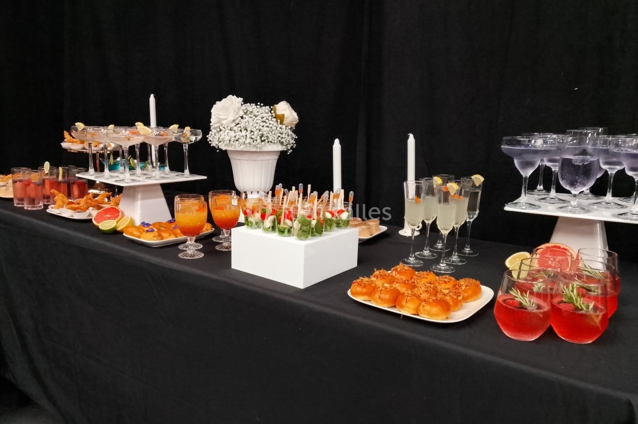 Buffet élégant avec cocktails colorés, verrines, amuse-bouches et décorations florales sur une nappe noire.
