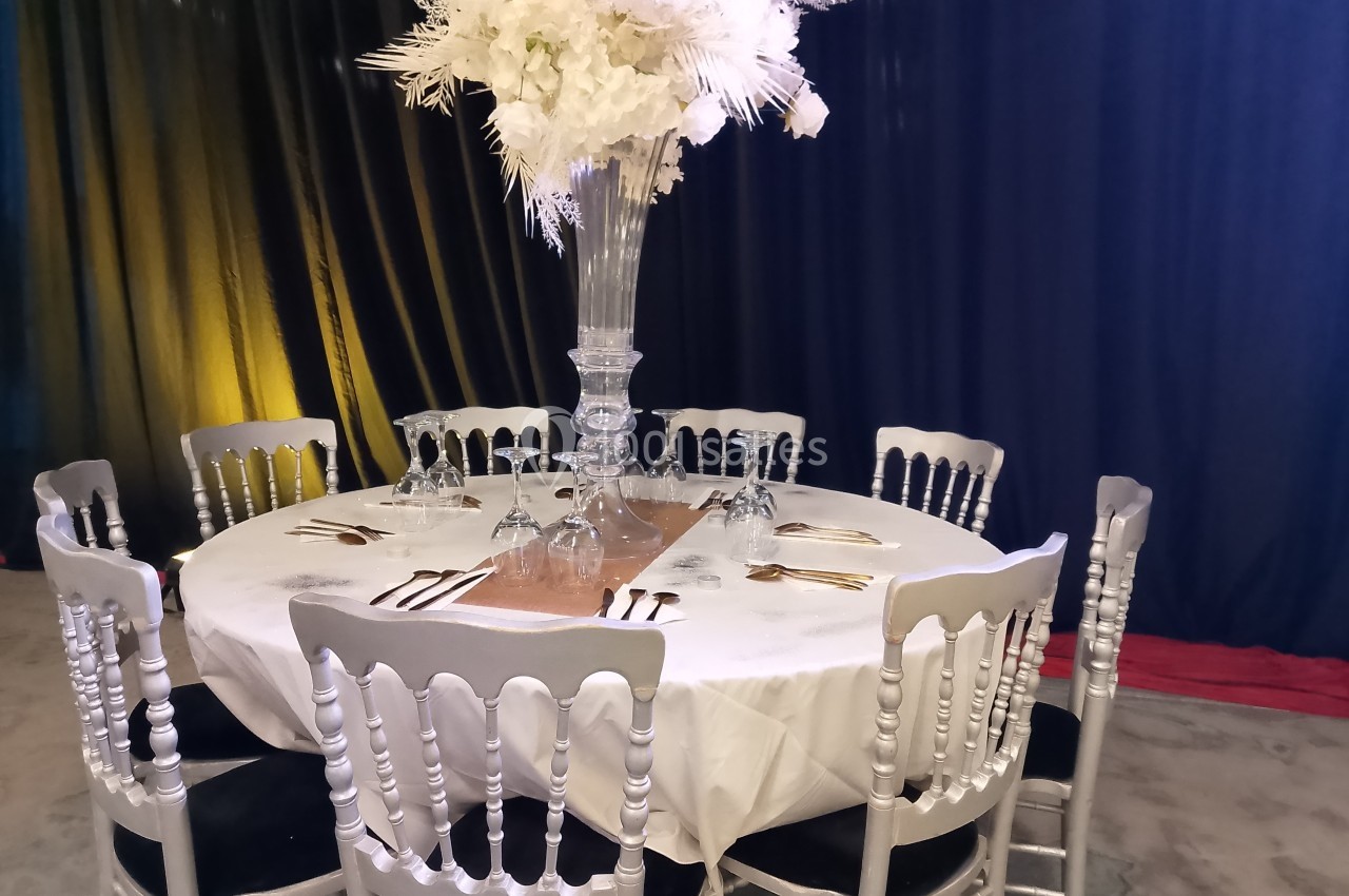 Table ronde dressée avec une nappe blanche, couverts dorés, verres et un grand centre de table floral blanc.