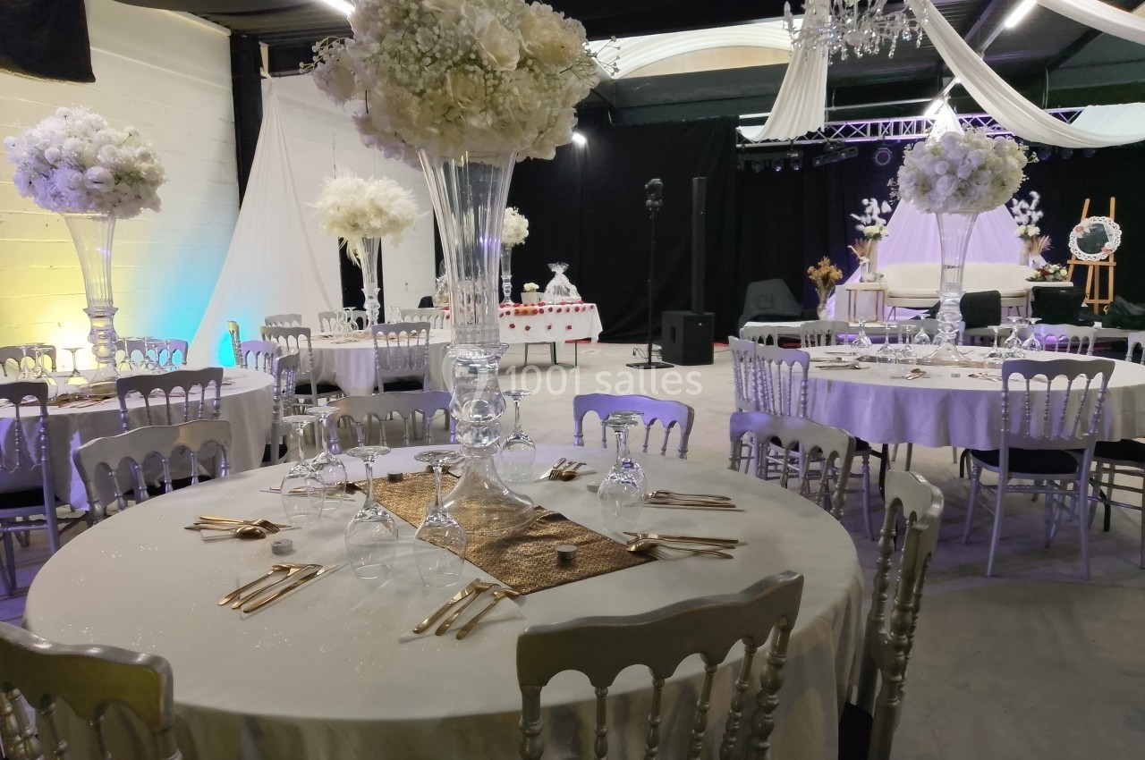 Salle de réception décorée avec des tables rondes, nappes blanches, centres de table fleuris et éclairage tamisé.