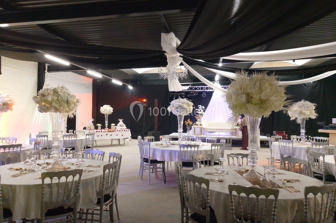 Salle de réception décorée avec des tables rondes, nappes blanches, chaises argentées et arrangements floraux élégants.