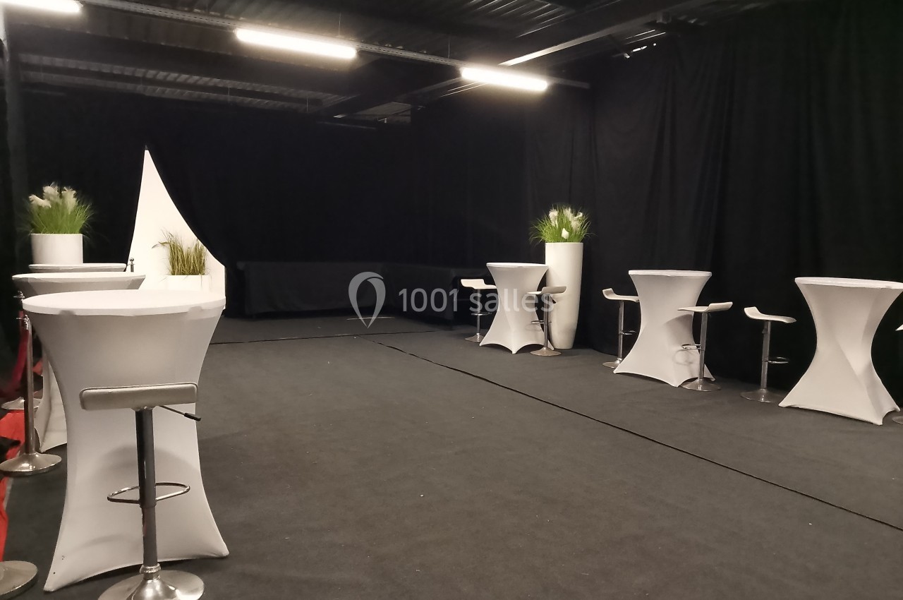 Salle aménagée avec des tables hautes recouvertes de nappes blanches, tabourets et plantes décoratives.