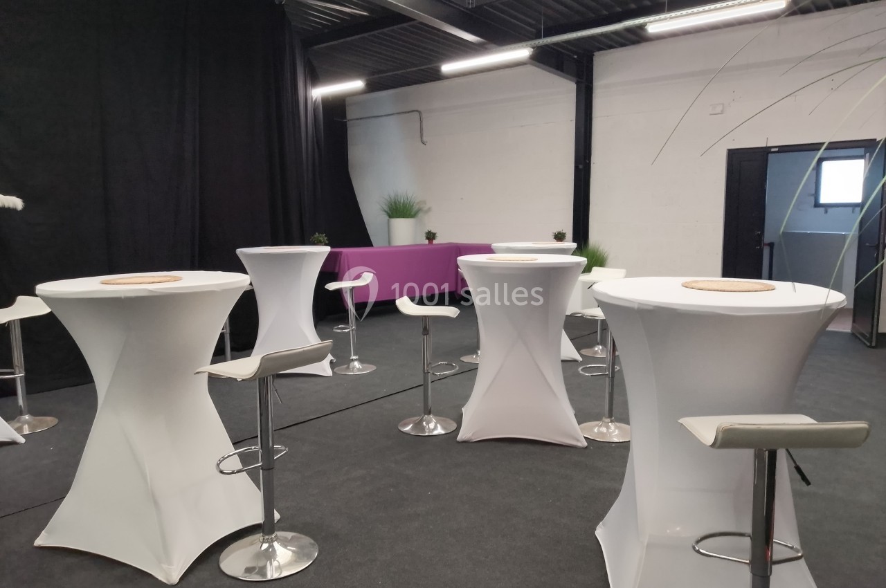 Salle aménagée avec des tables hautes blanches, des tabourets et une banquette violette, éclairée par des néons.