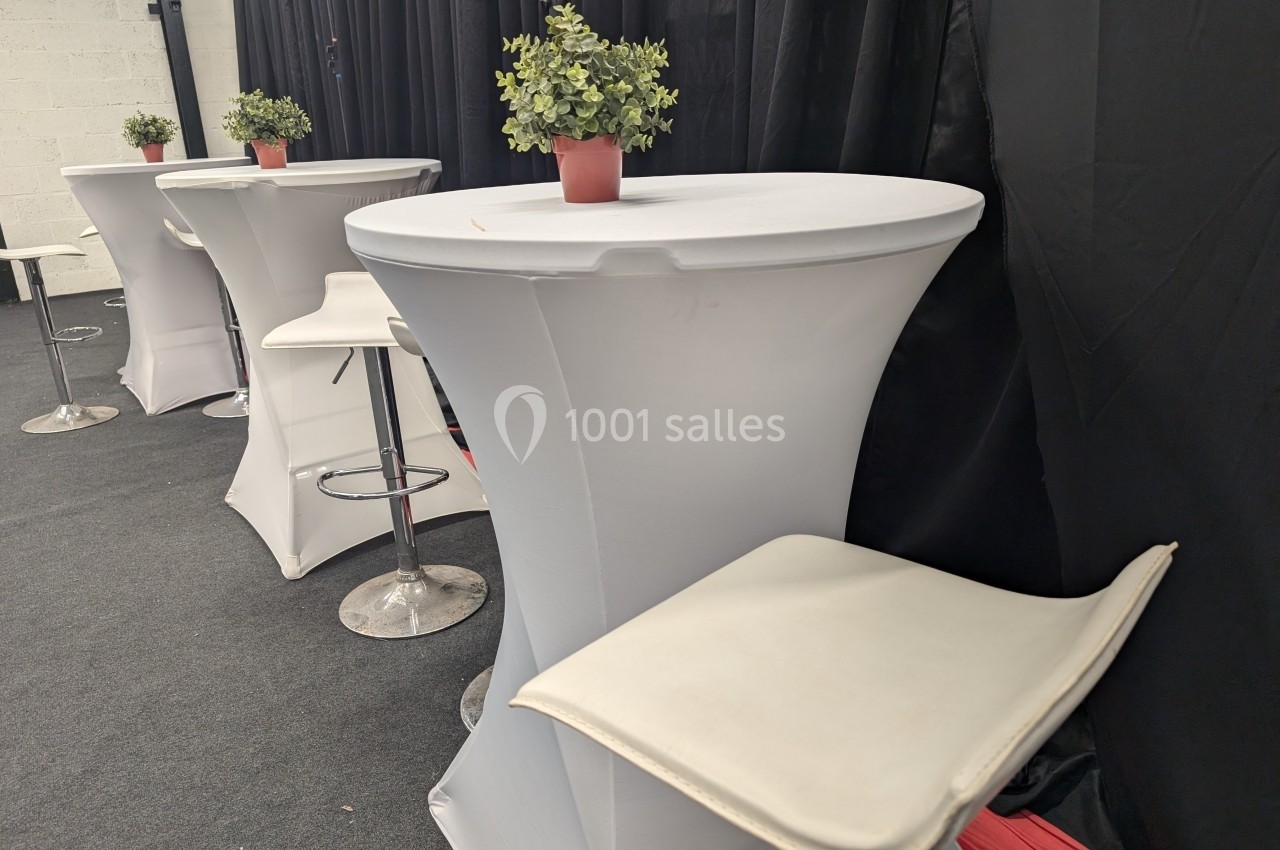 Tables hautes avec nappes blanches, tabourets et petites plantes en pot dans un espace intérieur avec rideaux noirs.
