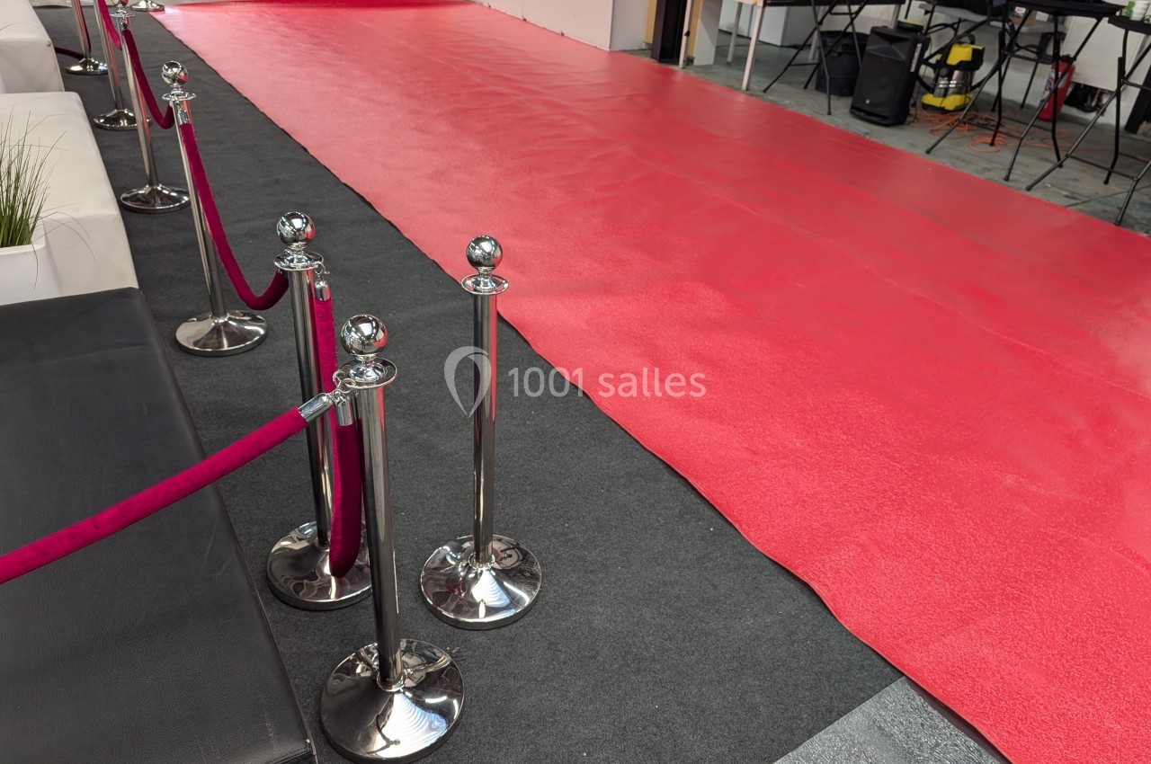 Tapis rouge déployé avec des poteaux et cordons, entouré de mobilier et d'équipements en arrière-plan.