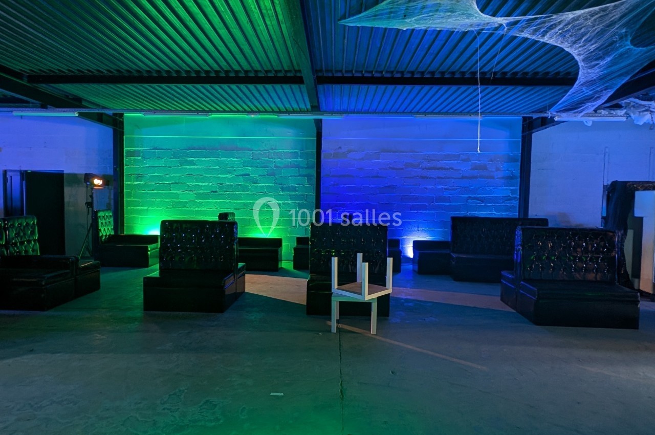Salle industrielle avec banquettes noires, éclairage coloré vert et bleu, et une chaise blanche au centre.