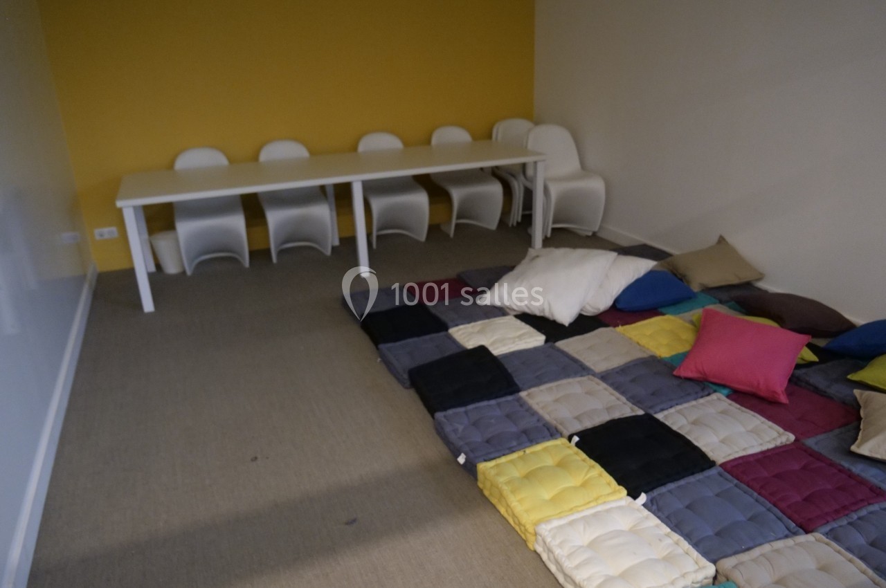 Salle avec un tapis de coussins colorés au sol, une table blanche et des chaises alignées contre un mur jaune.