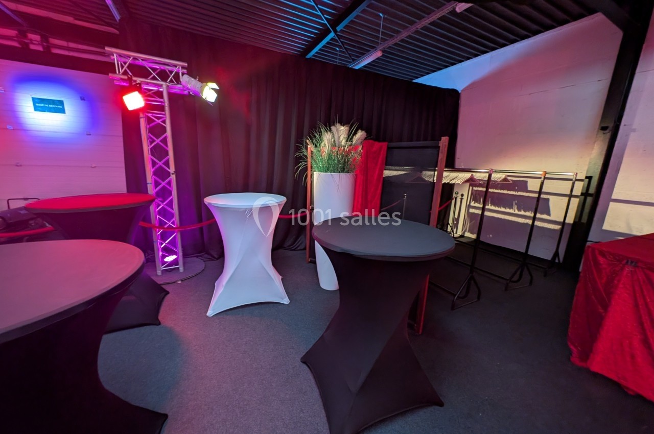 Salle aménagée avec des tables hautes recouvertes de housses, éclairage coloré et décorations végétales.