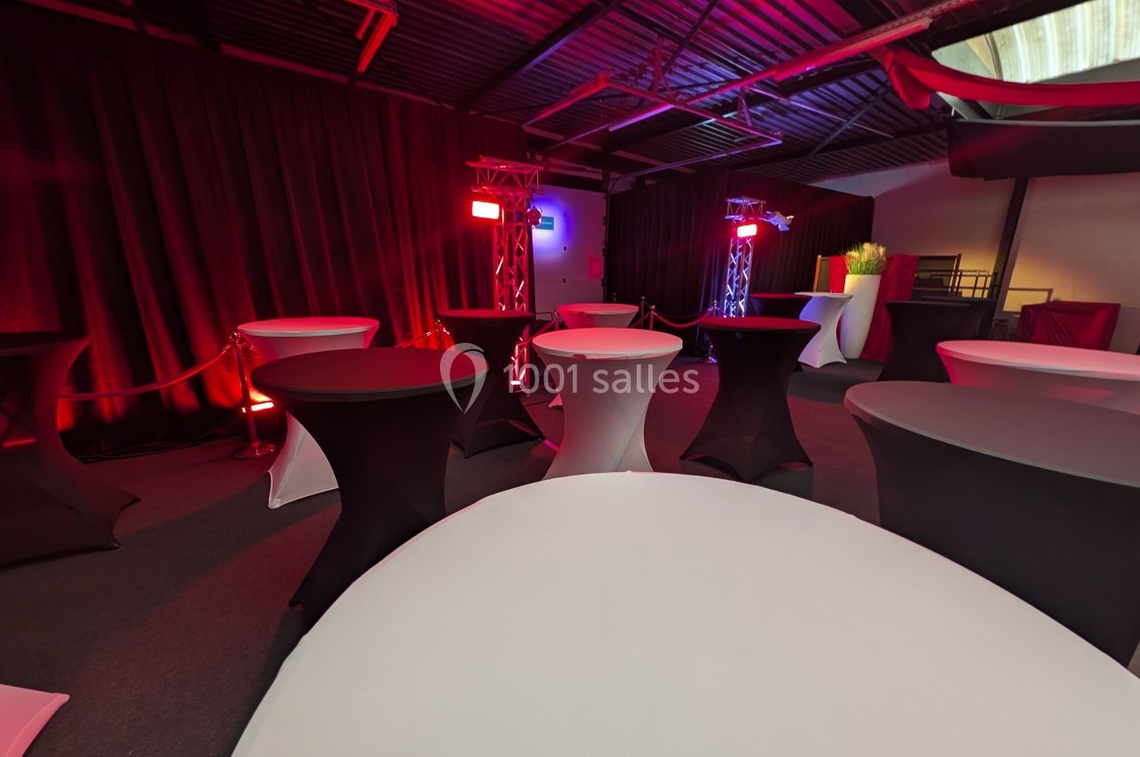 Salle avec des tables hautes recouvertes de nappes noires et blanches, éclairée par des lumières rouges et violettes.