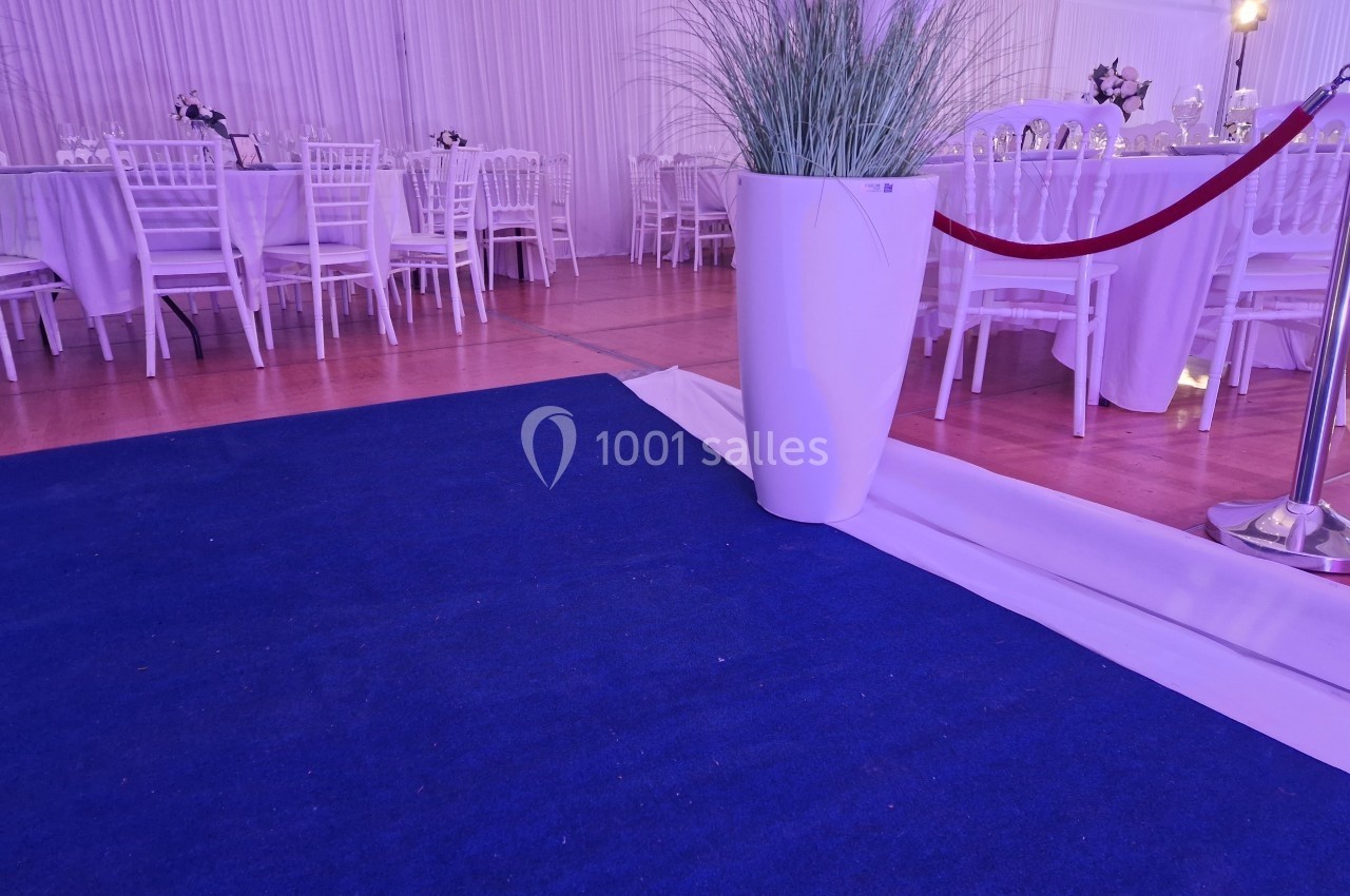 Tapis bleu menant à une salle de réception avec des tables dressées, décorée de plantes et de barrières rouges.