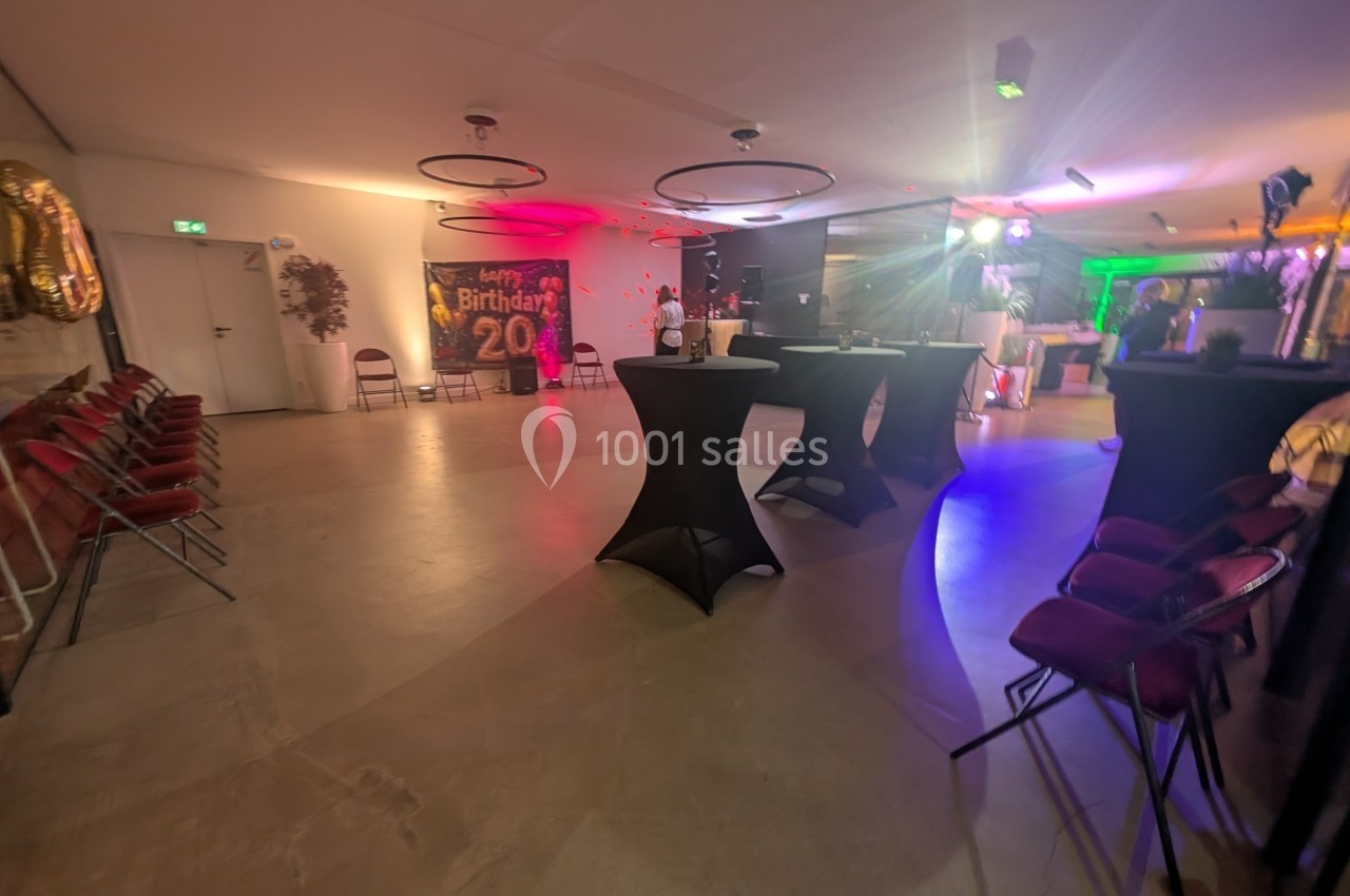 Salle décorée pour une fête avec des tables hautes, des chaises pliantes et une bannière d'anniversaire colorée.