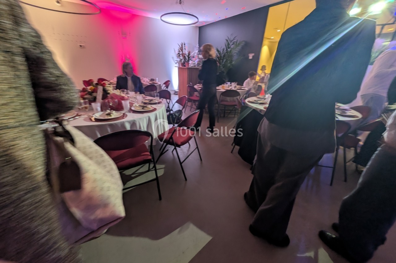 Salle de réception avec tables dressées, invités debout et assis, éclairage tamisé et ambiance festive.