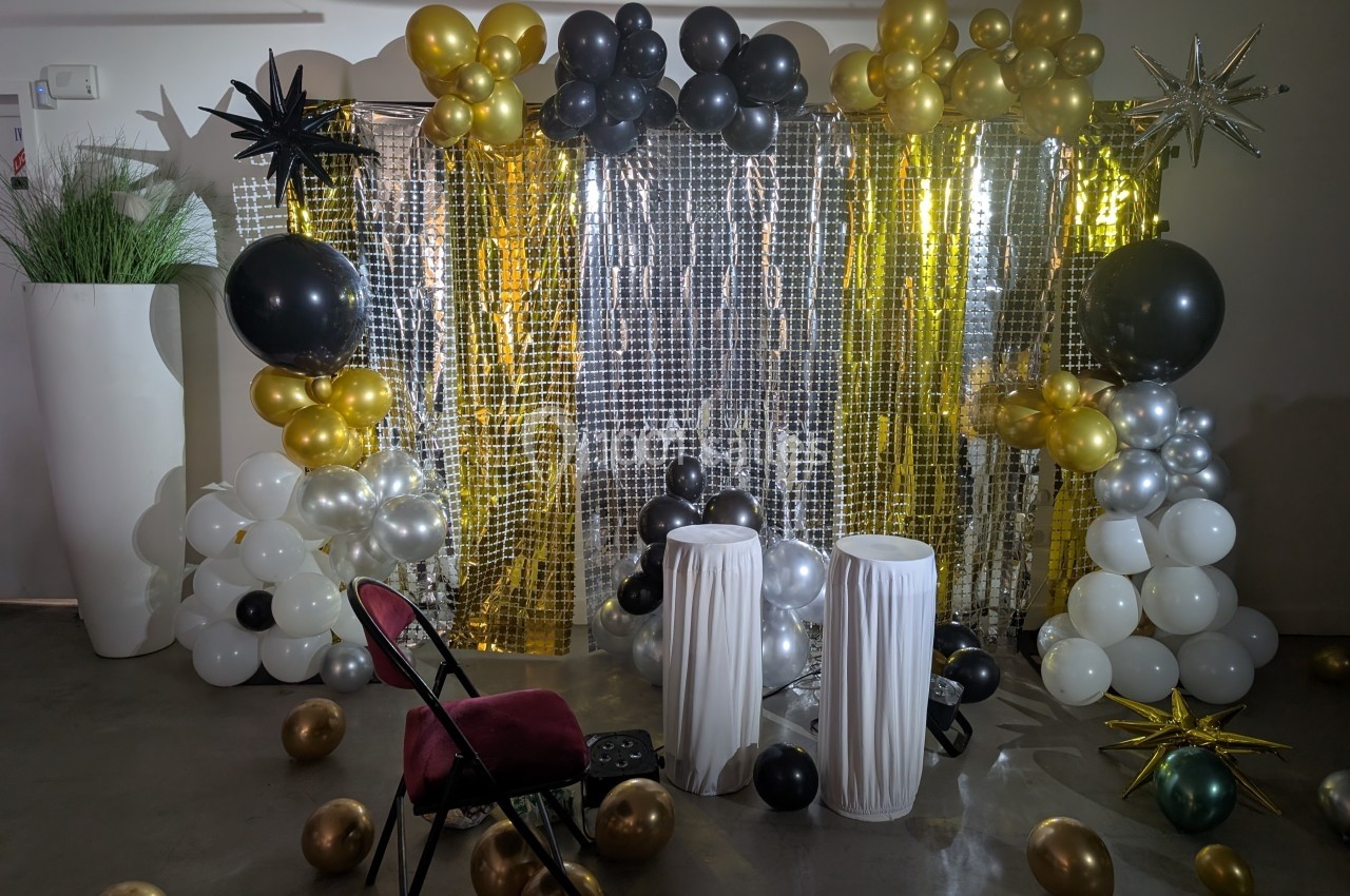 Décor festif avec ballons noirs, dorés, argentés et blancs, fond scintillant et deux tabourets blancs devant.