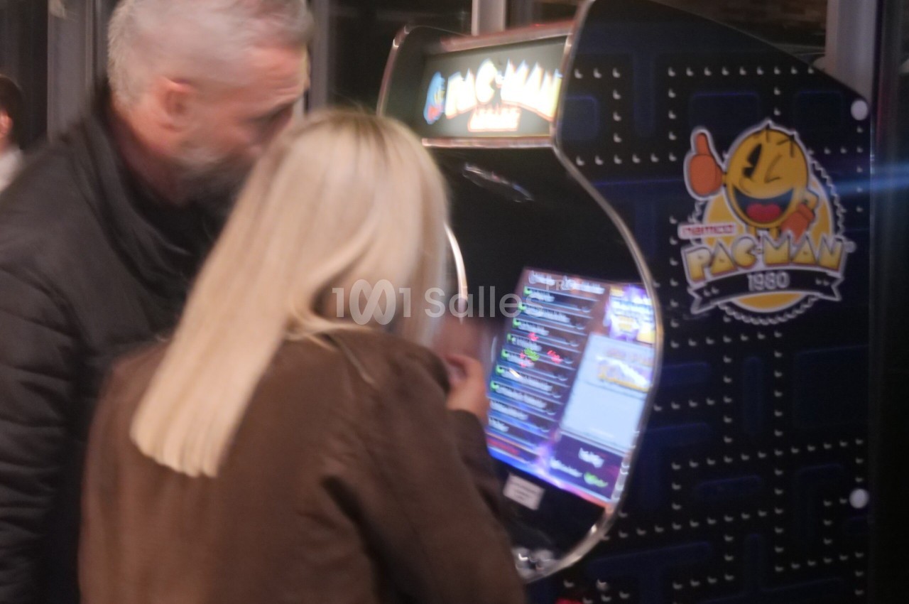 Un homme et une femme jouent sur une borne d'arcade rétro Pac-Man dans un espace intérieur.