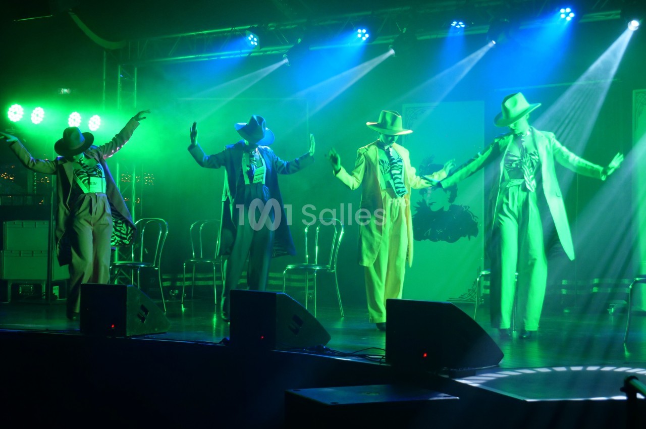 Quatre danseurs en costumes et chapeaux exécutent une chorégraphie sur une scène éclairée de lumières vertes.