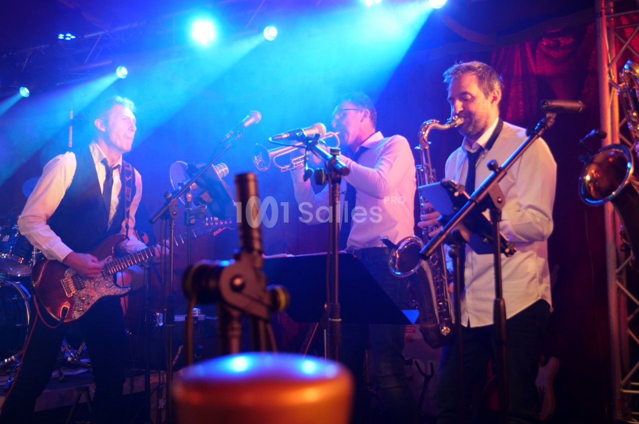 Musiciens jouant de la guitare, de la trompette et du saxophone sur une scène éclairée par des lumières bleues.