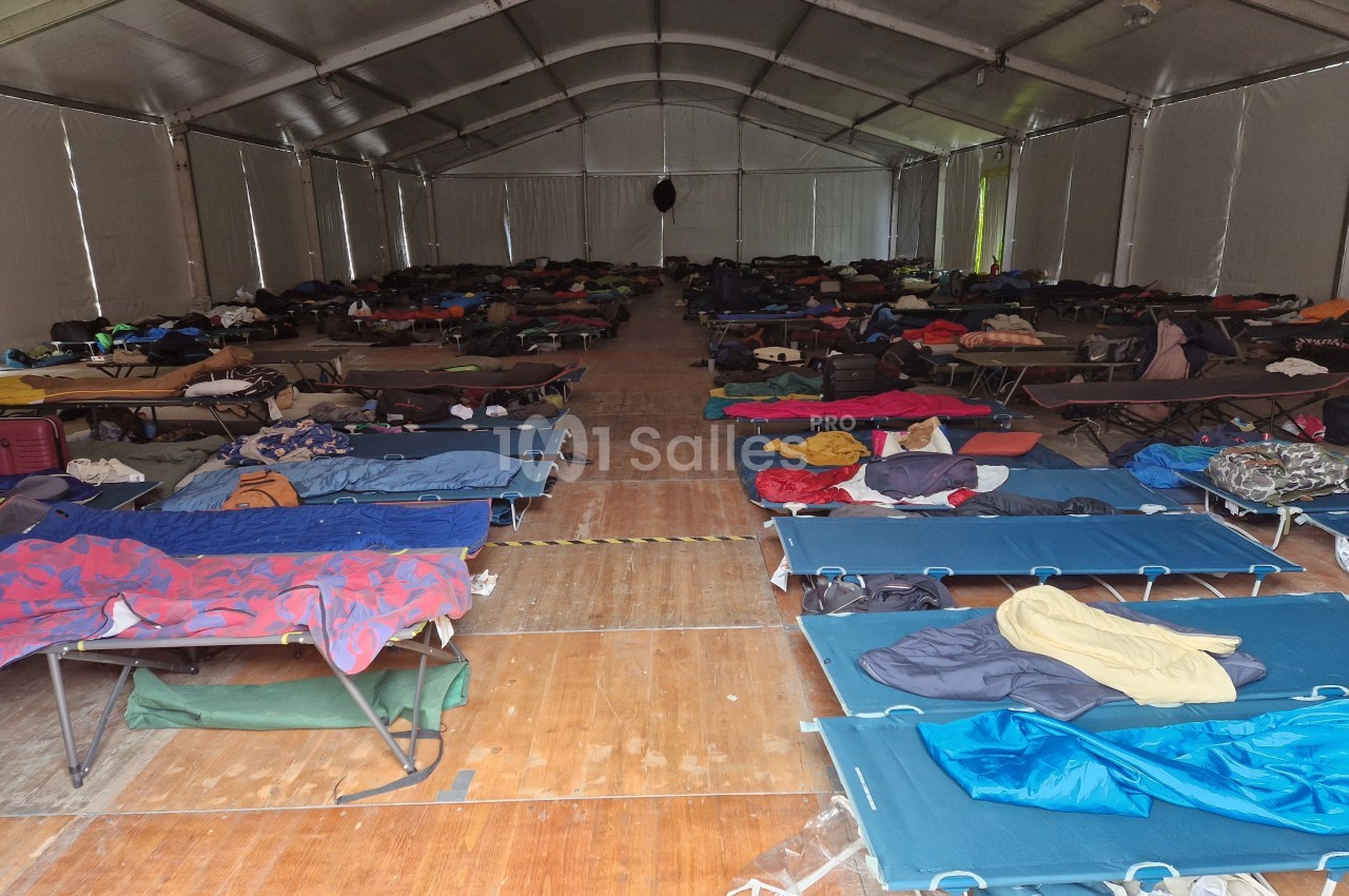 Intérieur d'un grand dortoir sous tente avec des rangées de lits de camp occupés par des affaires personnelles.