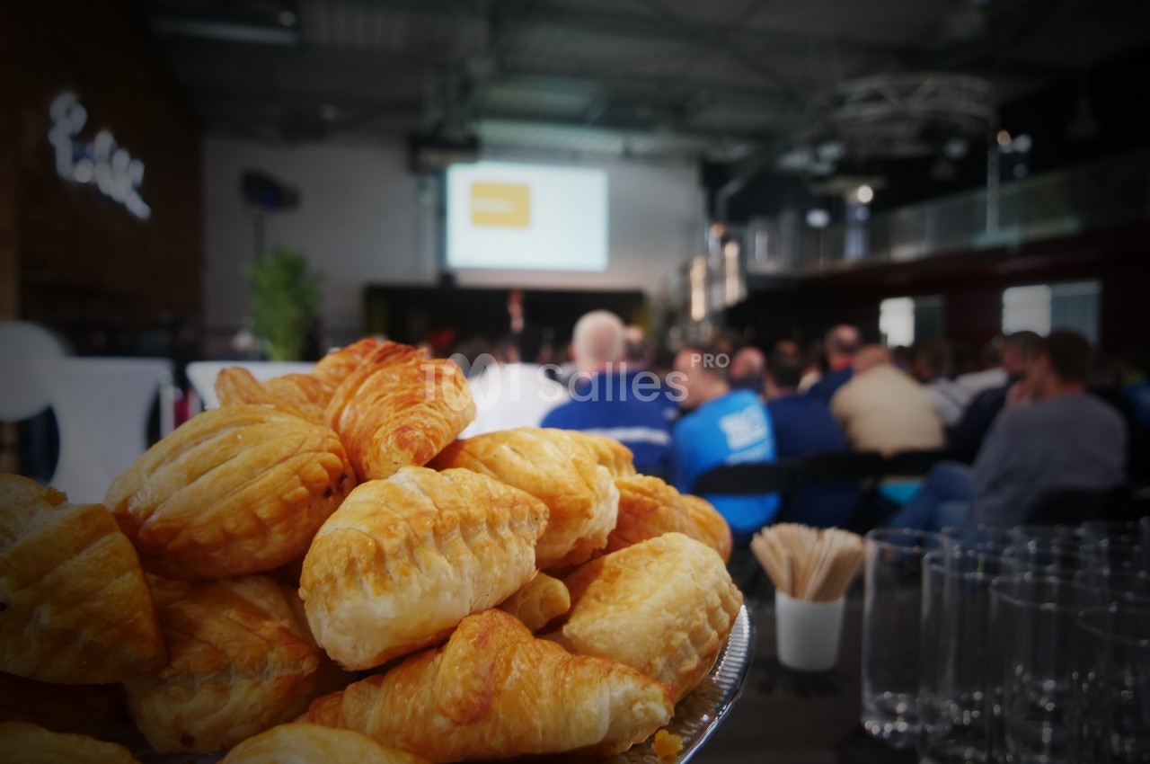 Assiette de viennoiseries au premier plan avec un public assis et un écran de présentation en arrière-plan.