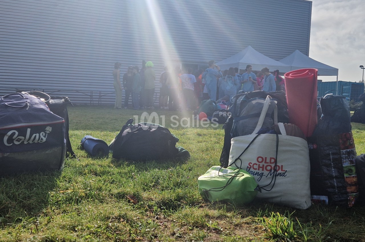 Des sacs et du matériel posés sur une pelouse, avec un groupe de personnes rassemblées près d'une tente blanche.