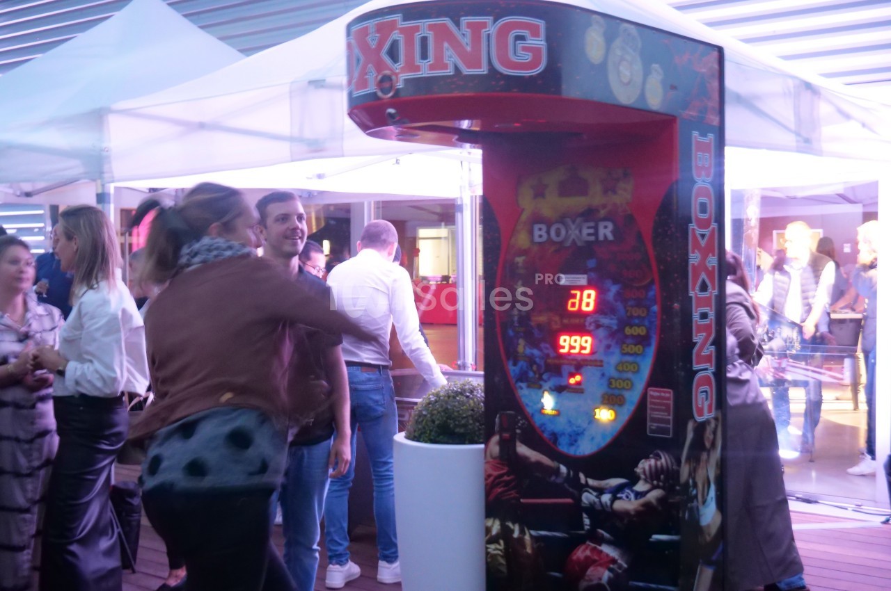 Machine de boxe électronique dans un espace extérieur animé, entourée de personnes discutant et observant.