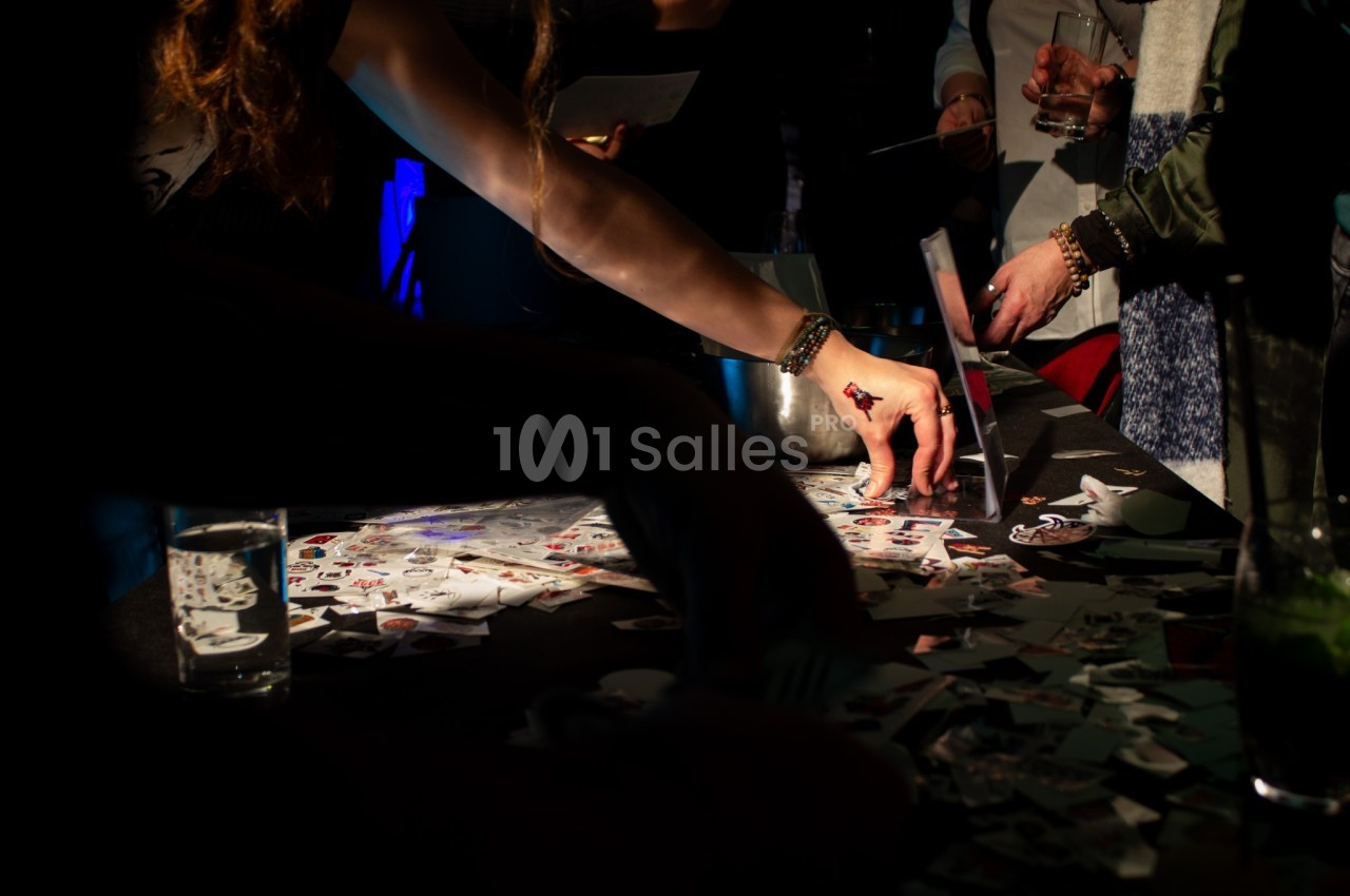 Des personnes choisissent des autocollants sur une table sombre éclairée, entourée de verres et de papiers.