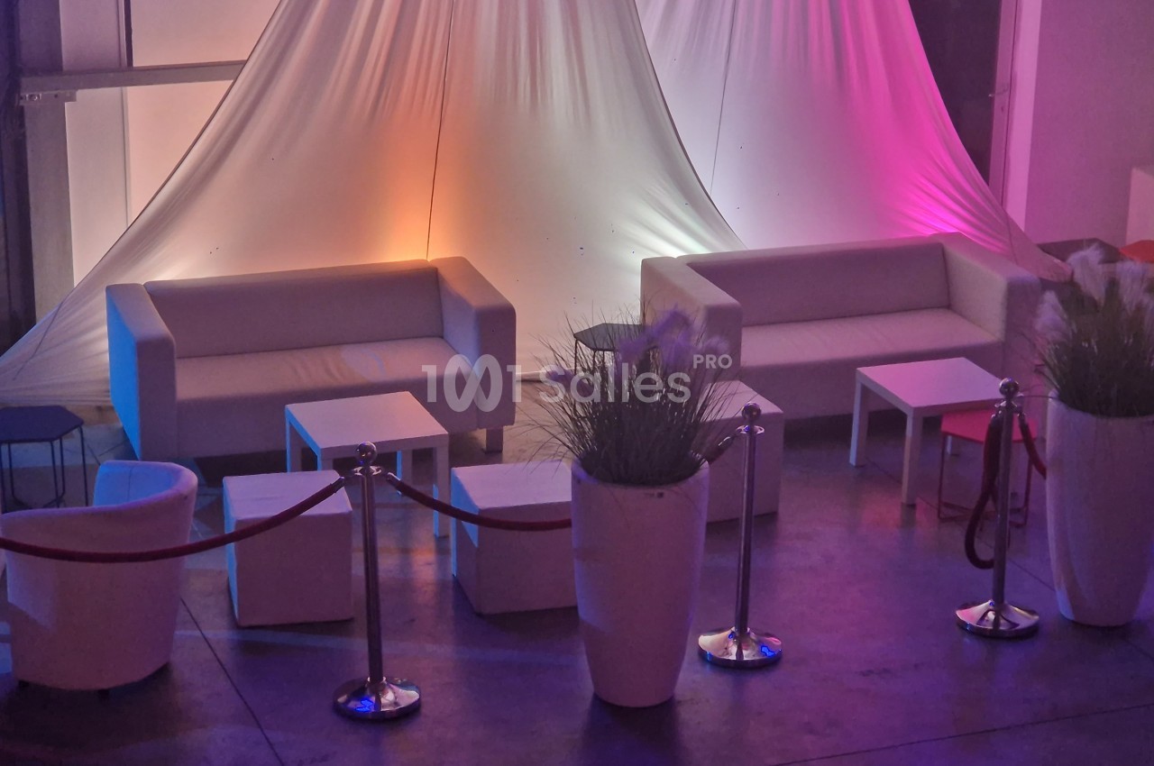 Deux canapés blancs entourés de tables basses et de plantes, dans un espace éclairé par des lumières colorées.