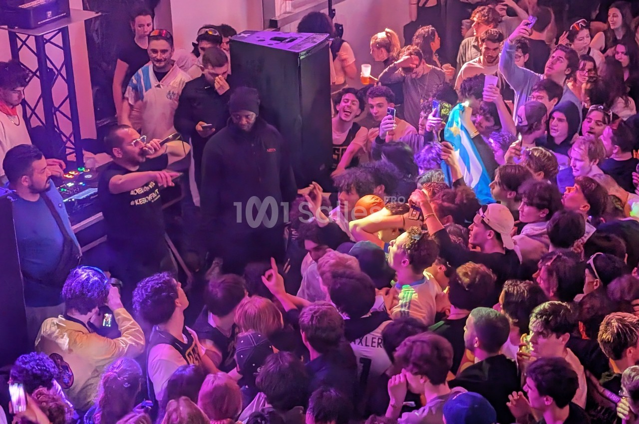 Foule rassemblée dans une salle de concert, avec des artistes sur scène et des spectateurs animés sous des lumières colorées.