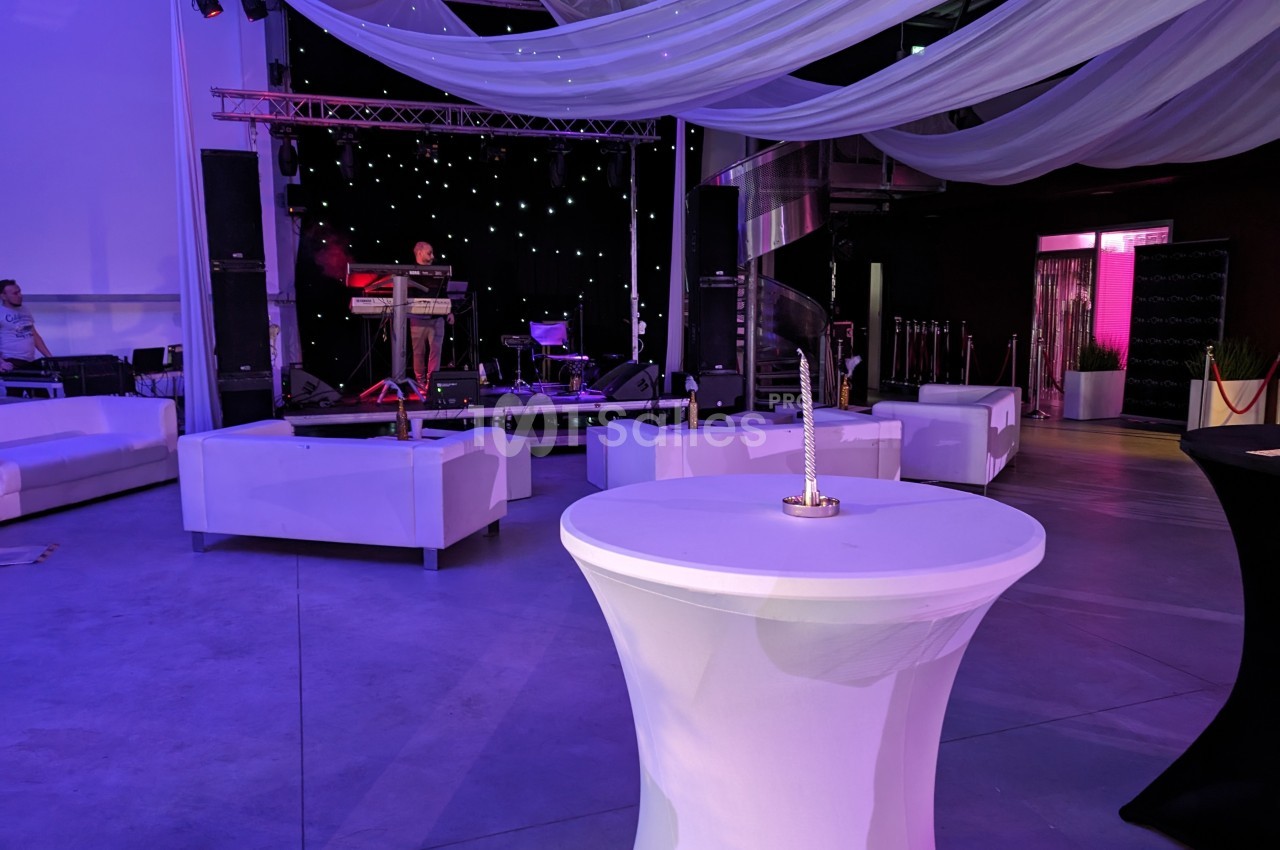 Salle événementielle avec scène éclairée, tables hautes et canapés blancs, décorée de voilages suspendus.