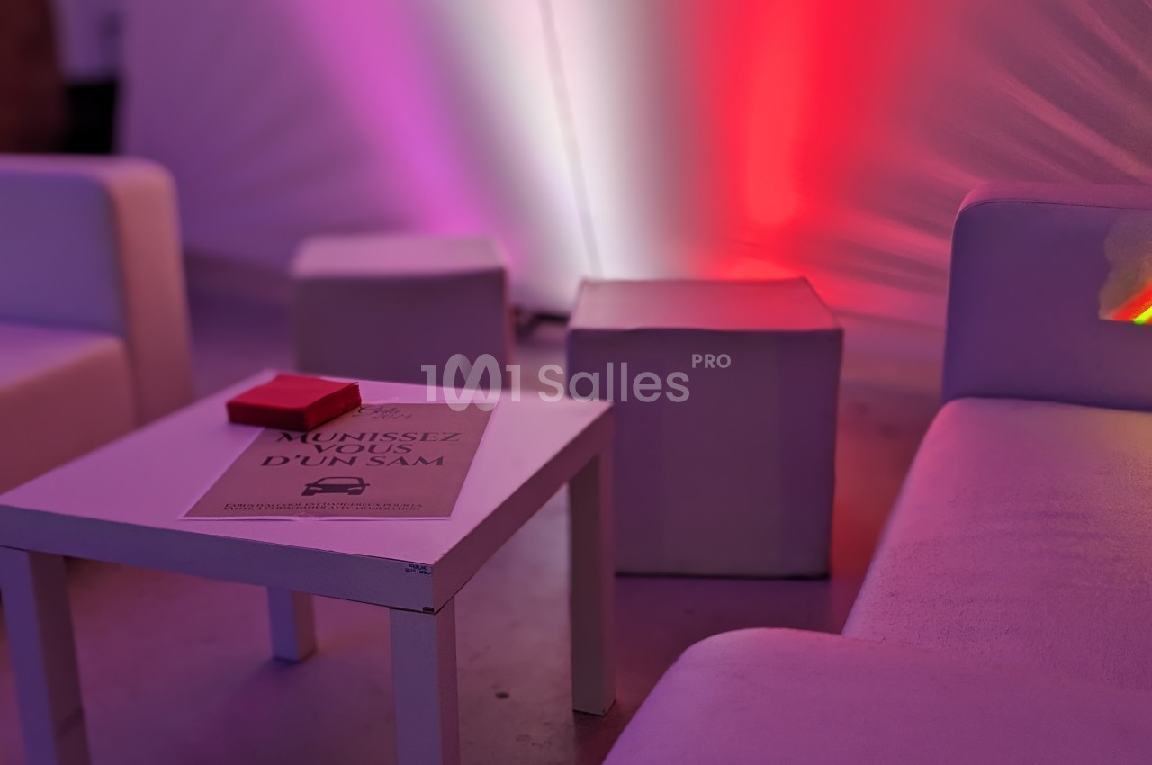 Table basse blanche avec un dépliant posé dessus, entourée de fauteuils et poufs dans une ambiance lumineuse colorée.