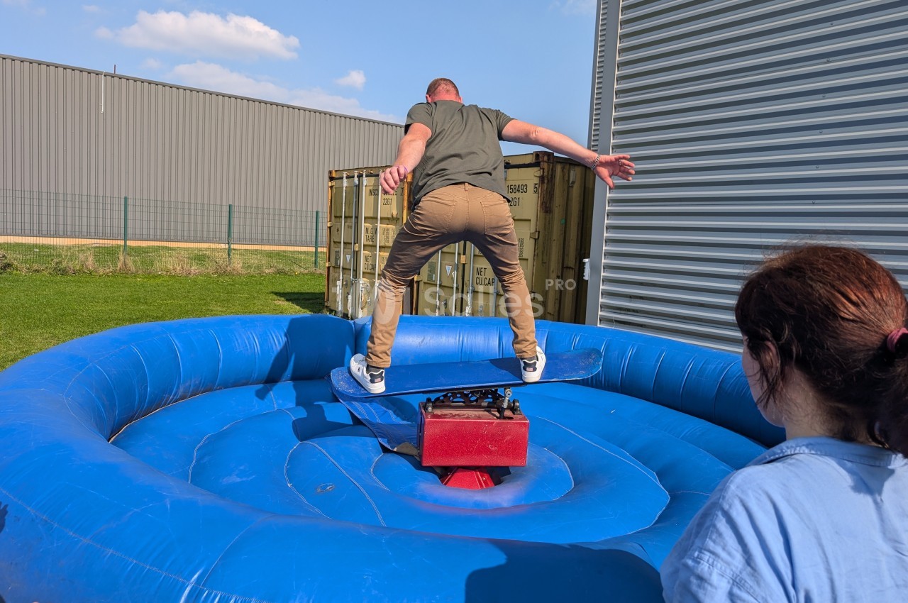 Un homme tente de garder l'équilibre sur une planche mécanique installée sur une structure gonflable bleue.