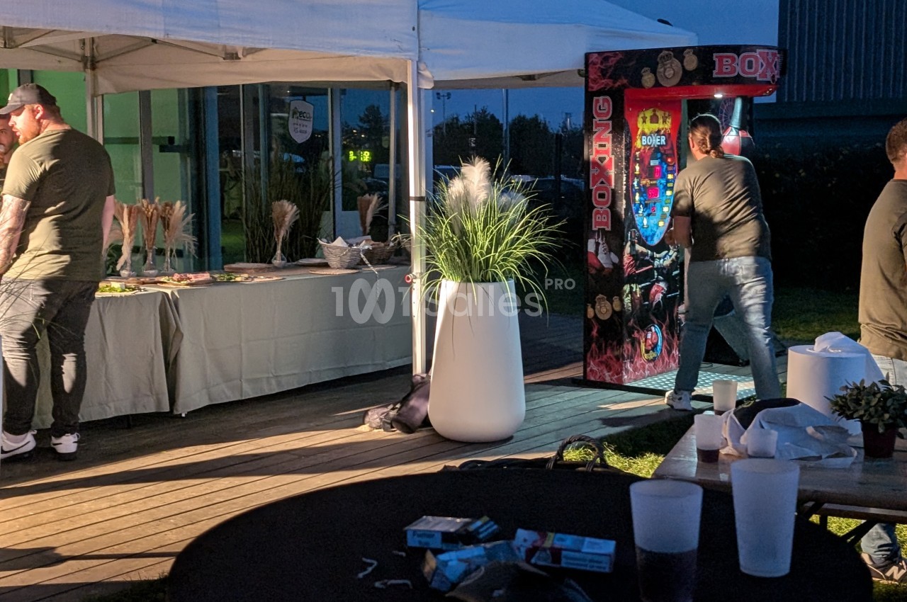 Un homme frappe une machine de boxe sous une tente lors d'un événement en plein air, avec des tables et décorations autour.