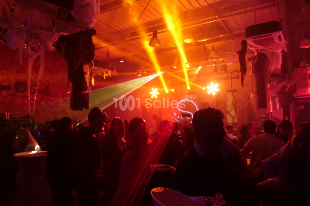 Salle de fête animée avec des lumières colorées, des invités debout et une scène éclairée en arrière-plan.
