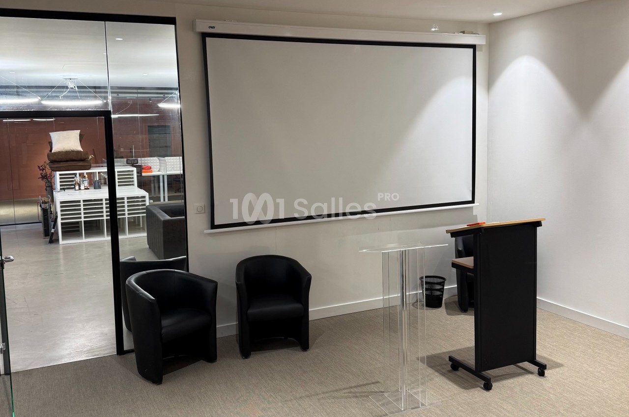 Salle de réunion avec écran de projection, pupitre transparent, deux fauteuils noirs et vue sur un espace adjacent.