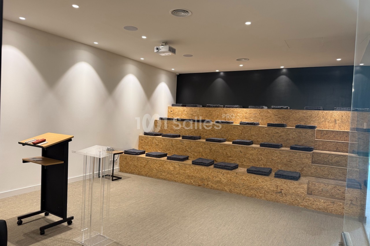 Salle de conférence avec gradins en bois, coussins noirs, pupitre en bois et plexiglas, éclairage au plafond.