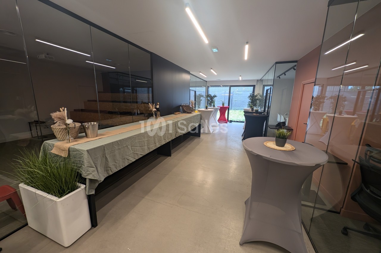 Salle moderne avec tables dressées pour un événement, décorée de plantes et éclairée par des baies vitrées.