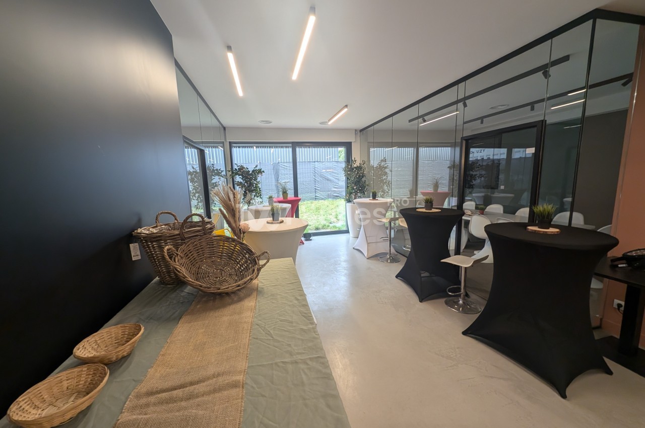 Salle lumineuse avec tables hautes, nappes noires, paniers en osier sur une table et vue sur un jardin extérieur.