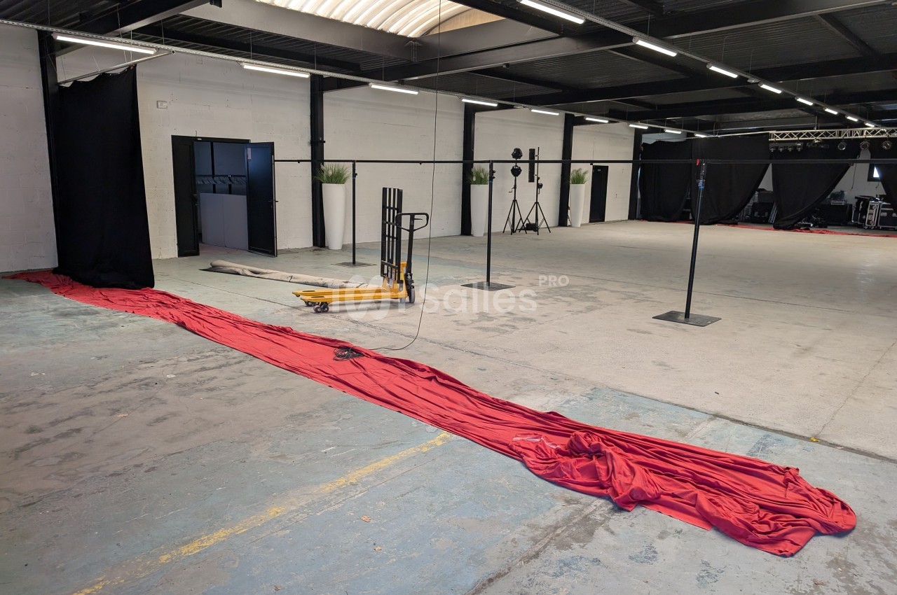 Grand espace intérieur industriel avec sol en béton, rideau rouge étendu au sol et équipements de studio en arrière-plan.