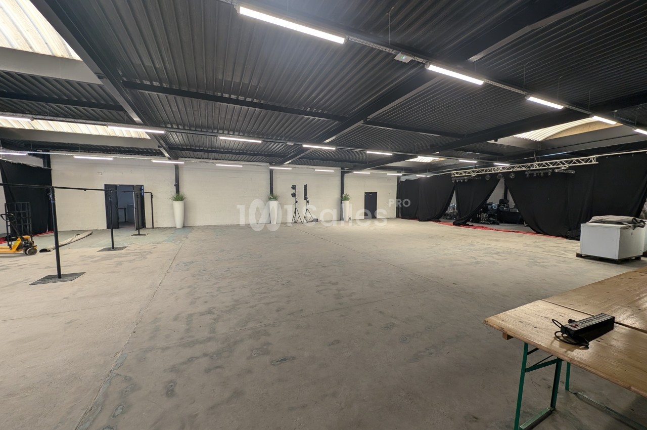 Grande salle vide avec sol en béton, plafond métallique, éclairage suspendu et scène équipée de rideaux noirs au fond.