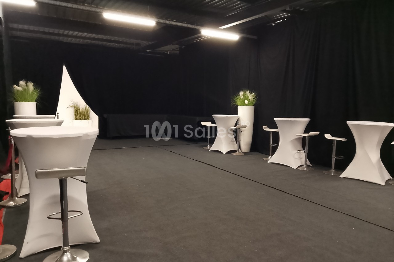 Salle aménagée avec des tables hautes recouvertes de nappes blanches, tabourets et plantes décoratives.