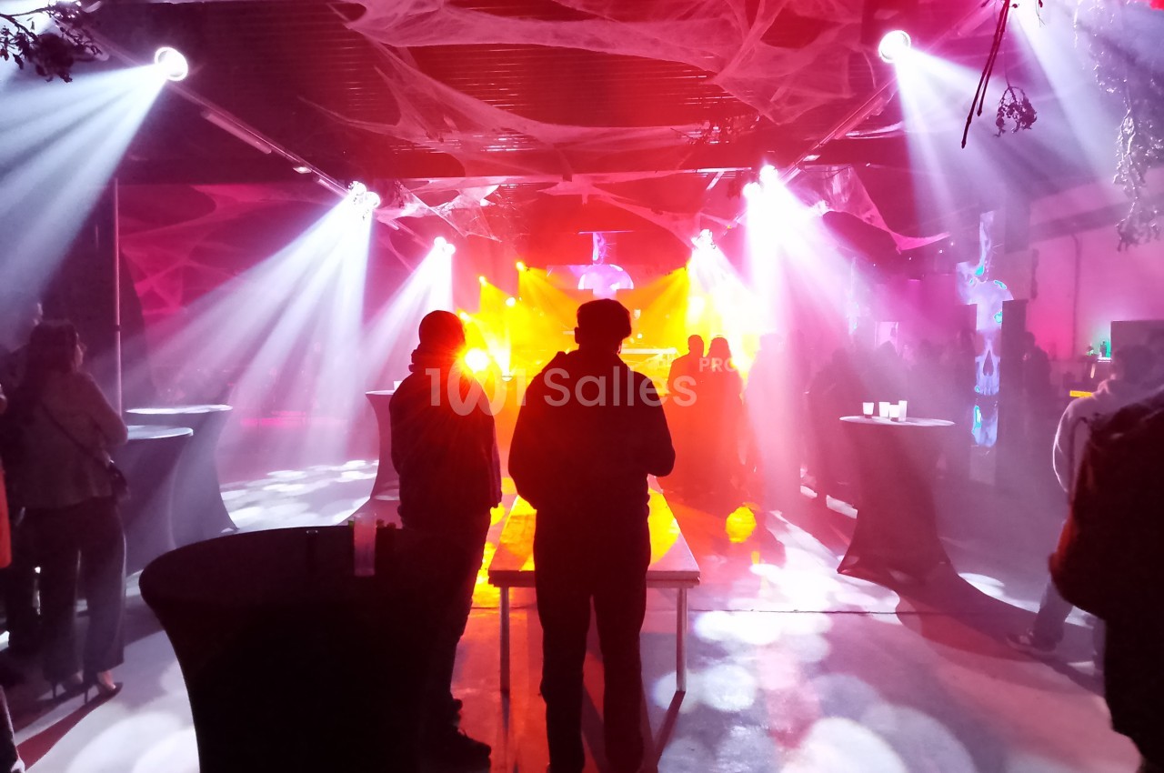 Salle sombre décorée pour une fête, éclairée par des lumières colorées avec des silhouettes de personnes debout.