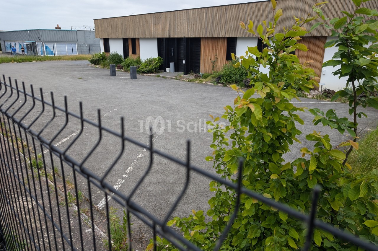 Bâtiment moderne avec façade en bois, parking vide et végétation, vu à travers une clôture métallique.