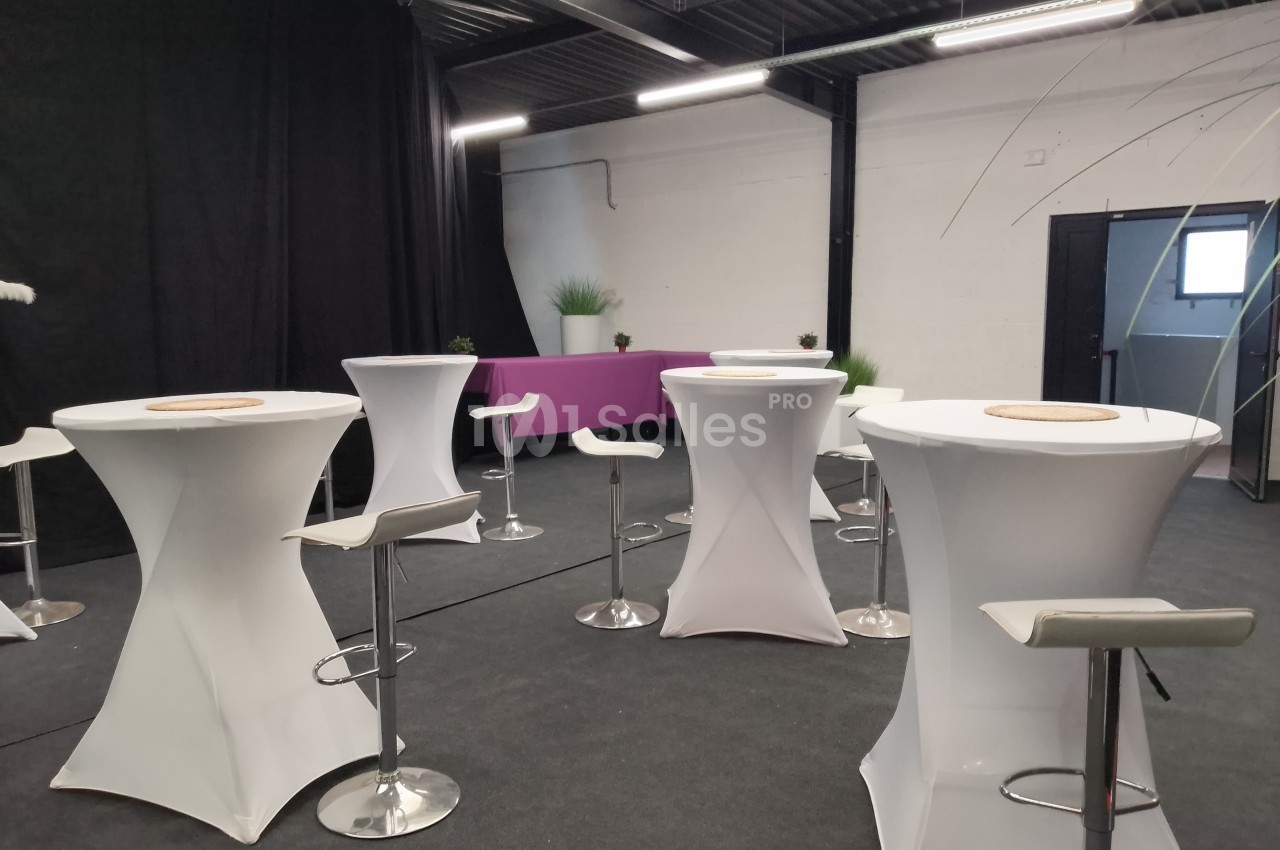 Salle aménagée avec des tables hautes blanches, des tabourets et une banquette violette, éclairée par des néons.
