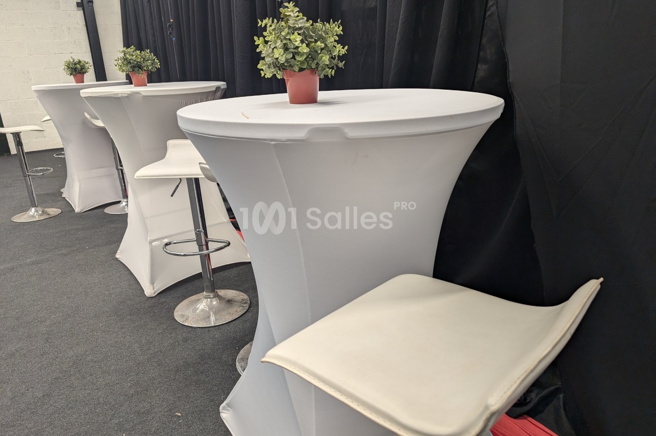 Tables hautes avec nappes blanches, tabourets et petites plantes en pot dans un espace intérieur avec rideaux noirs.
