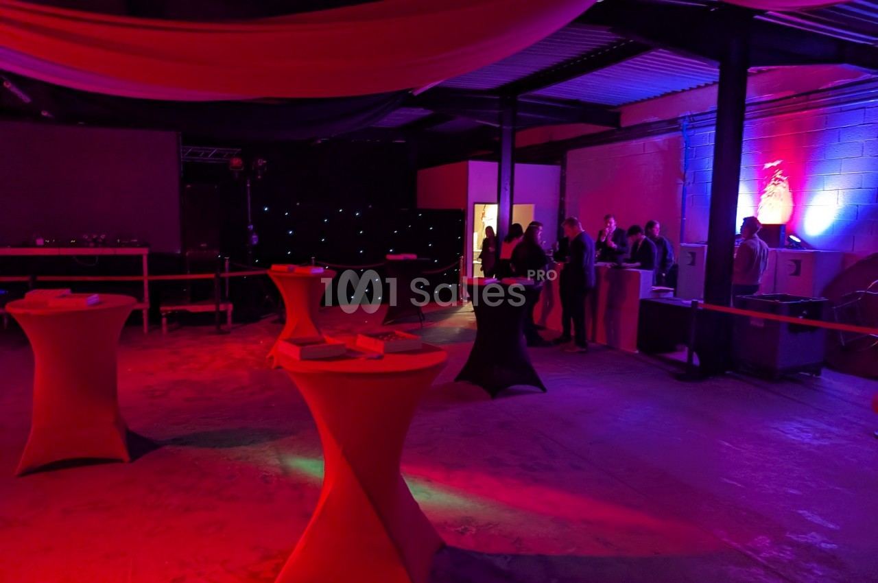 Salle sombre avec des tables hautes éclairées par des lumières rouges et bleues, et des personnes discutant près d'un…