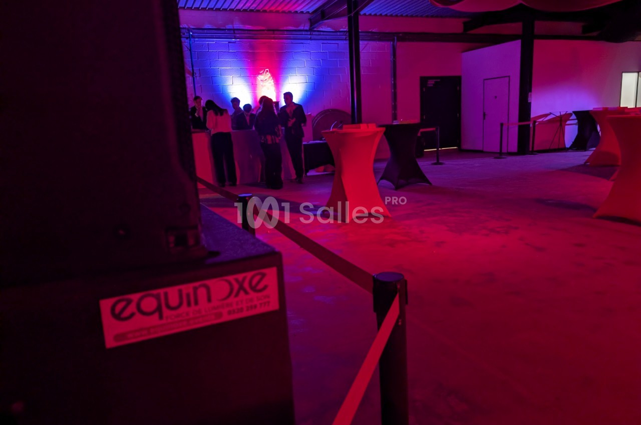 Salle sombre éclairée par des lumières rouges et bleues, avec des tables hautes et un groupe de personnes au fond.