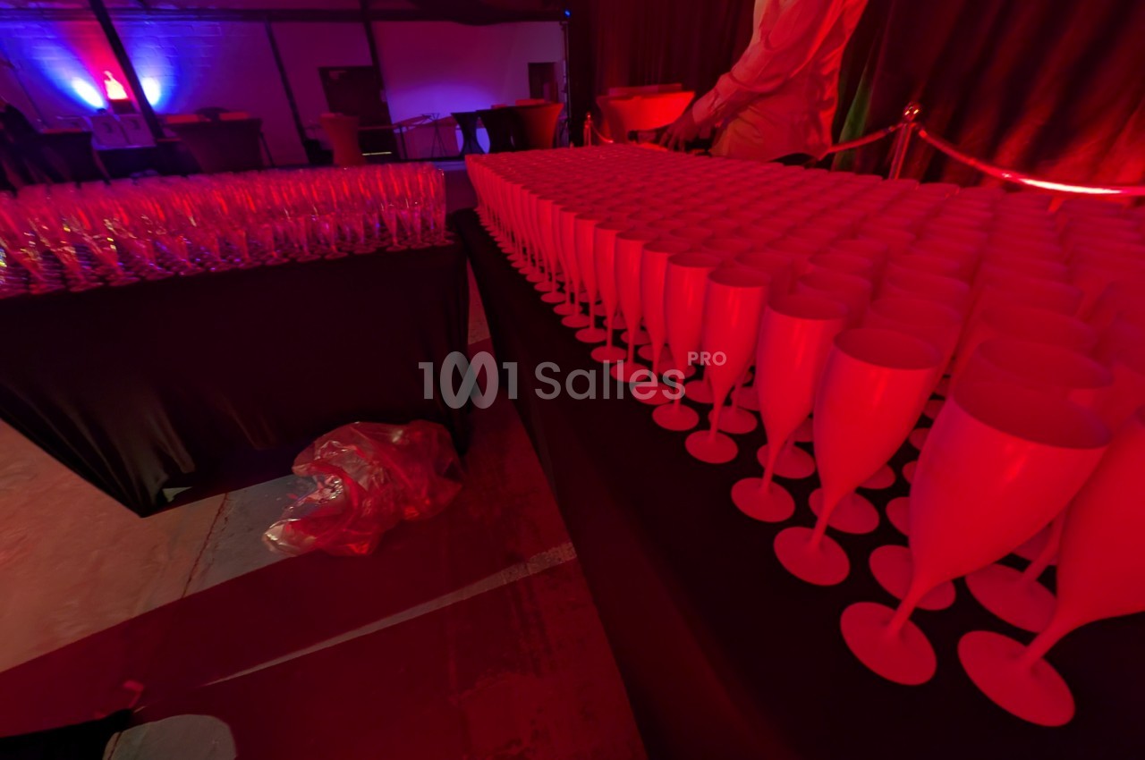 Rangées de verres à pied rouges alignés sur une table dans une salle éclairée par des lumières tamisées rouges et violettes.
