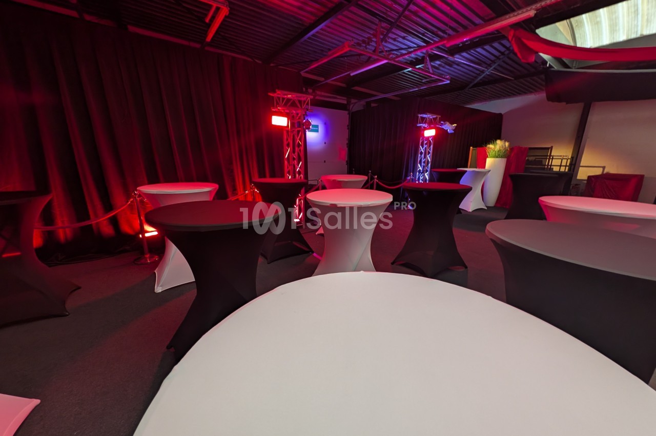Salle avec des tables hautes recouvertes de nappes noires et blanches, éclairée par des lumières rouges et violettes.