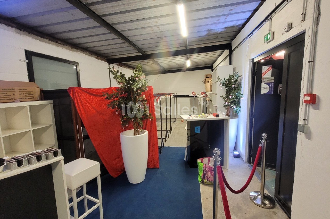 Espace intérieur avec sol bleu, plantes en pots, rideau rouge, mobilier blanc et noir, et barrière à cordon rouge.