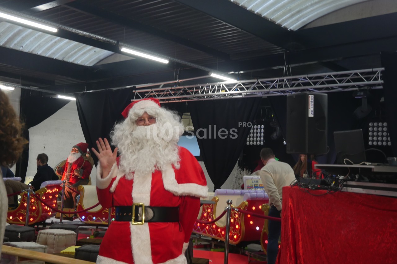Un Père Noël en costume rouge salue dans un décor festif avec guirlandes lumineuses et scène en arrière-plan.